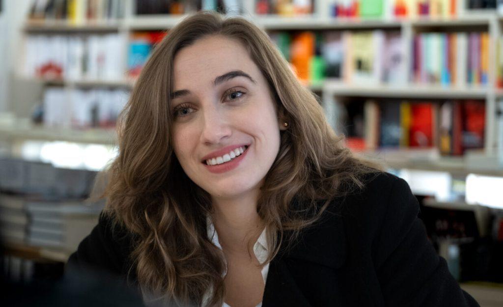 Mujer sonriente con cabello castaño ondulado, vistiendo chaqueta negra, frente a estantería con libros de fondo
