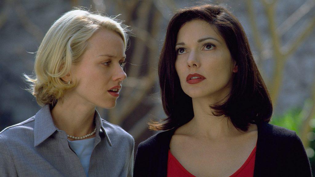 Dos mujeres conversan en un interior; una de cabello rubio claro viste de azul y la otra de cabello oscuro viste de rojo
