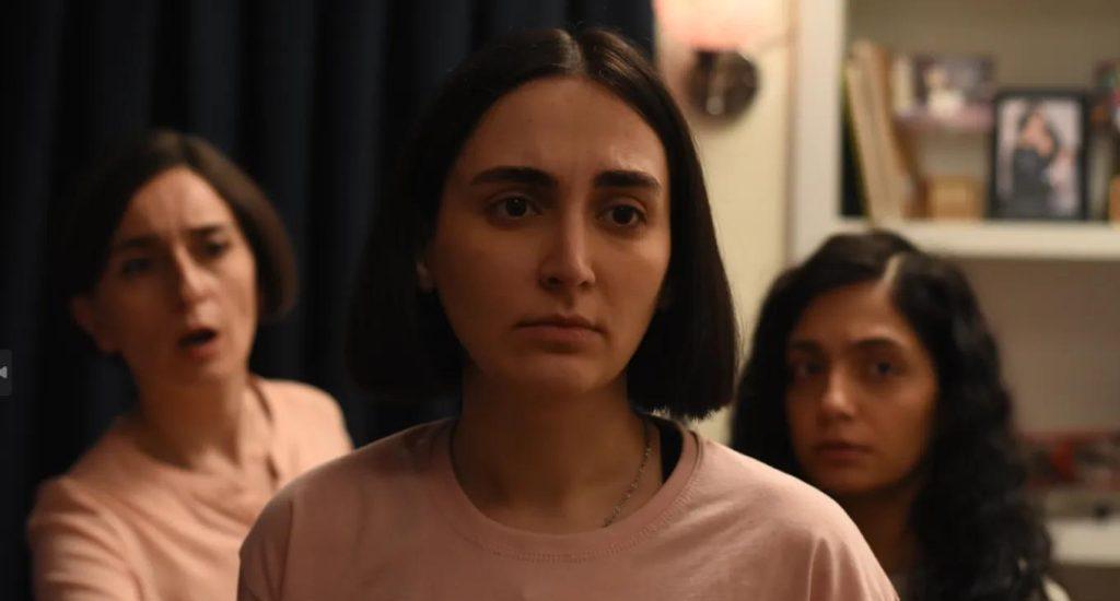 Tres mujeres en interior doméstico: una al frente con expresión seria, dos detrás observando, escena de tensión familiar