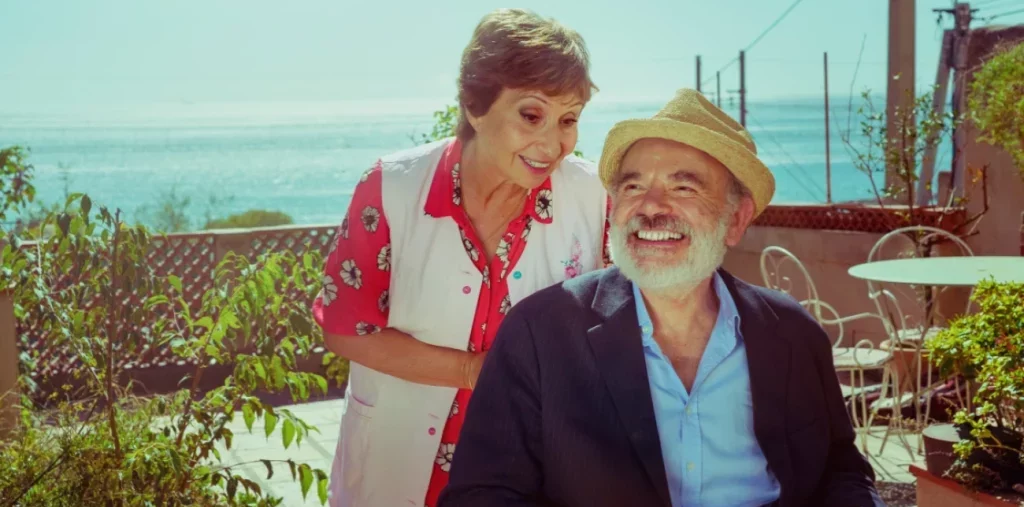 Dos personas en un jardín junto al mar; mujer de pie con camisa roja y blanca, hombre sentado con sombrero y camisa