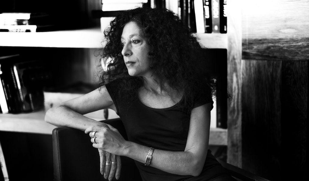 Leila Guerriero, escritora argentina de cabello rizado, sentada frente a estantería con libros, mirando hacia el lado
