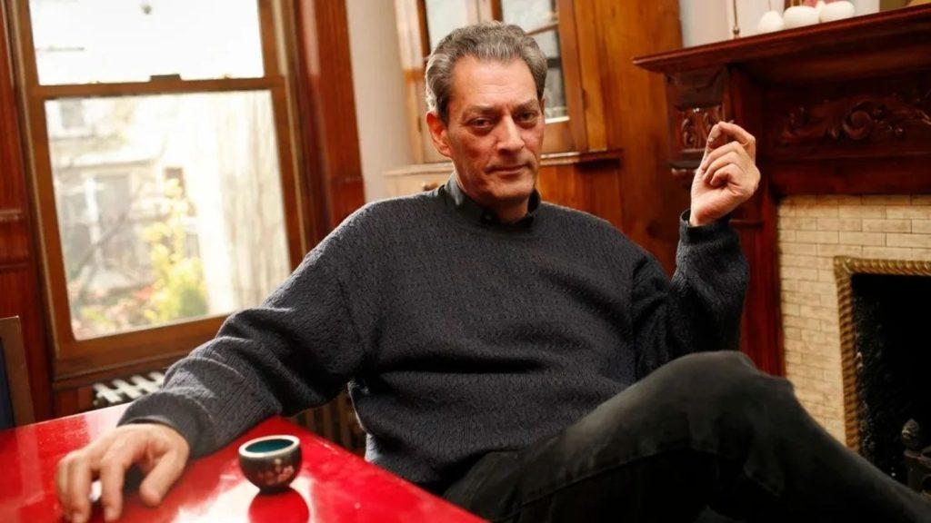 Paul Auster sentado en un sillón rojo, gesticulando mientras habla en una sala con chimenea y ventanas de madera