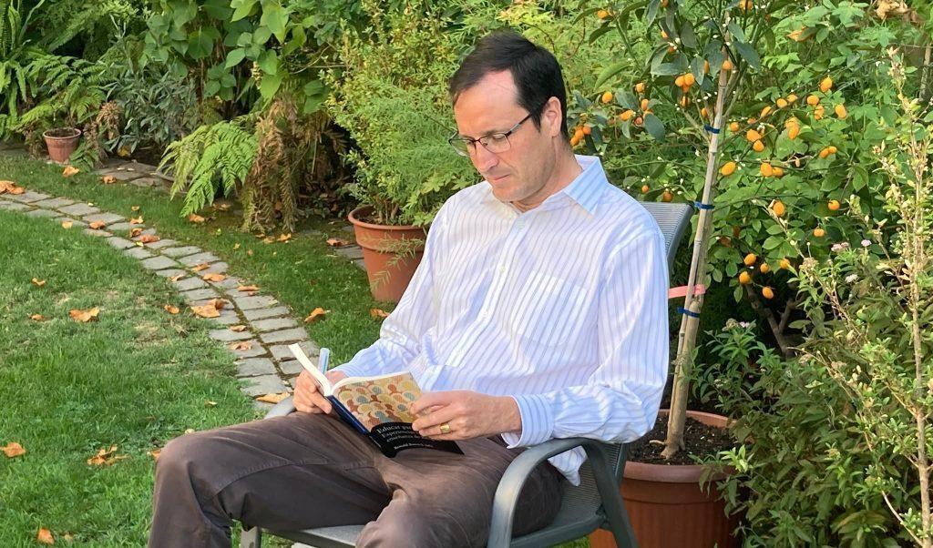 Ronald Brown sentado en jardín leyendo documento, rodeado de vegetación y plantas verdes
