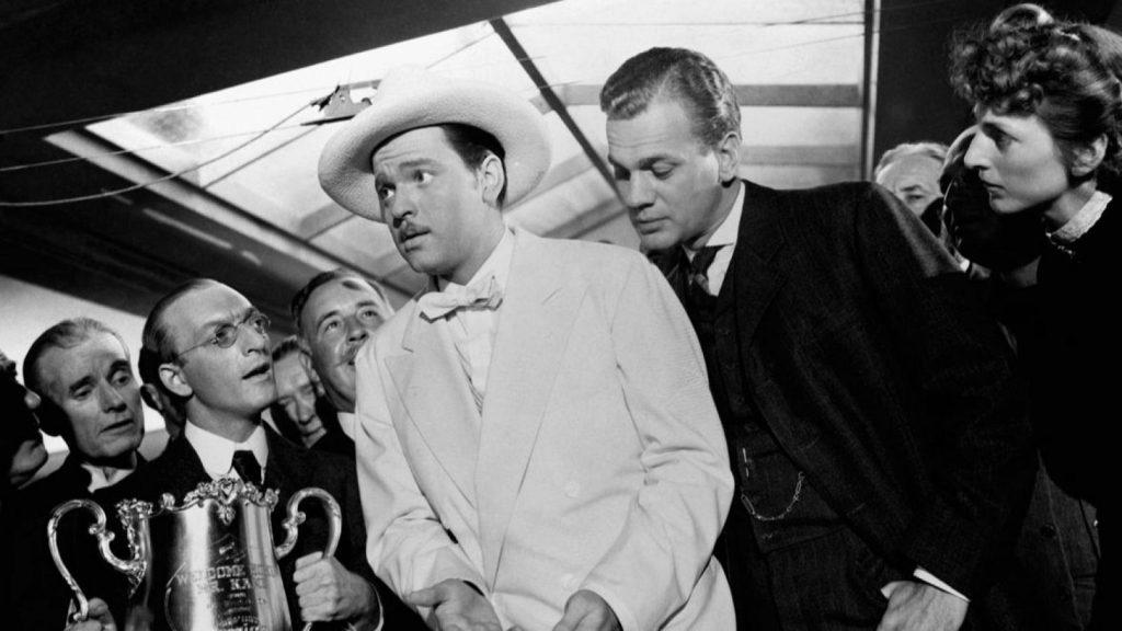 Orson Welles en traje blanco rodeado de hombres en escena de interrogatorio o confrontación cinematográfica