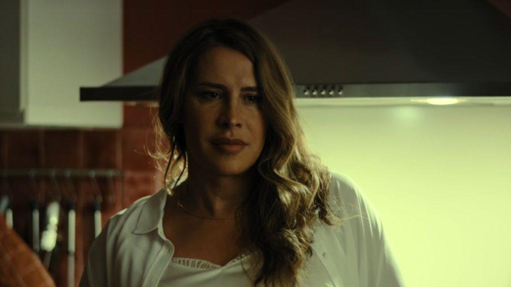 Mujer de cabello castaño ondulado en cocina iluminada, expresión seria, vistiendo blusa blanca clara