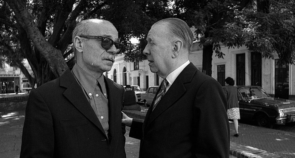 Jorge Luis Borges y Ernesto Sabato conversan en la calle, ambos de traje y gafas oscuras