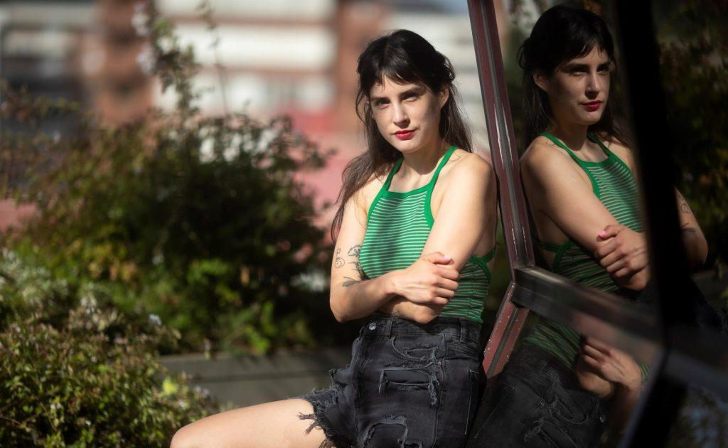 Paulina Flores, autora chilena, posa junto a una pared con reflejo, vistiendo top verde y pantalón gris oscuro