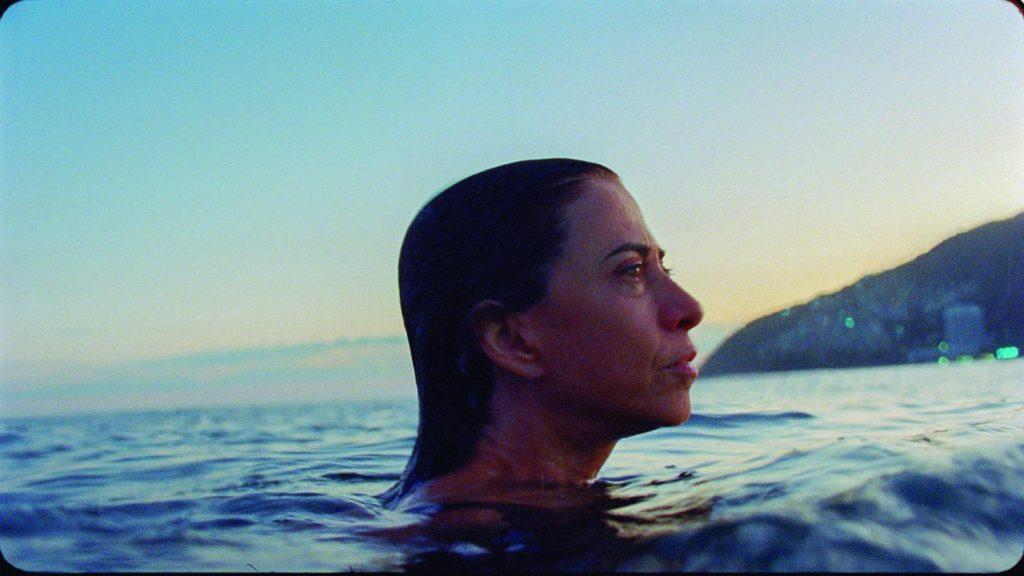 Mujer en el agua al atardecer, mirando hacia el horizonte con expresión reflexiva, montañas al fondo