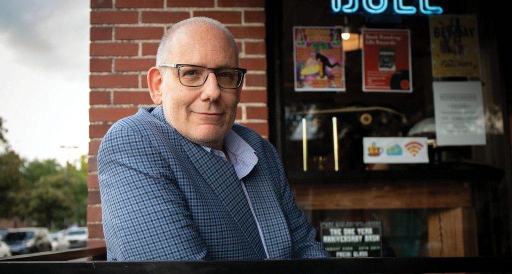 Hombre de mediana edad con gafas y camisa azul a cuadros sonriendo junto a estantería con libros
