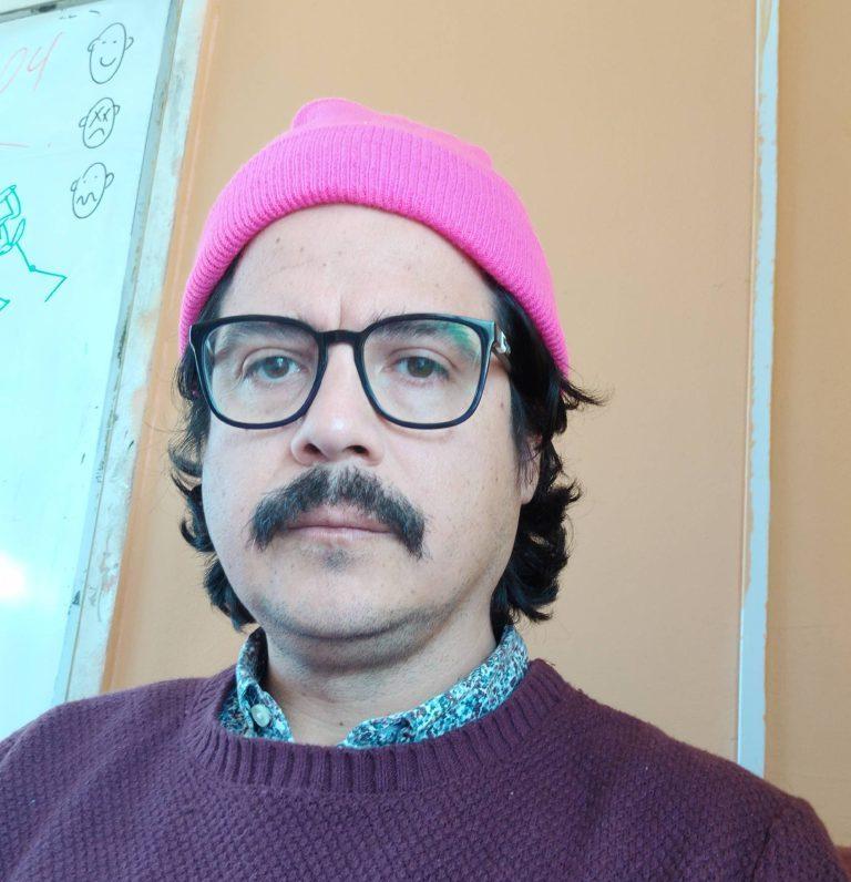 Hombre con gafas, bigote y gorro rosa, vistiendo suéter marrón sobre camisa azul, en interior con pizarra