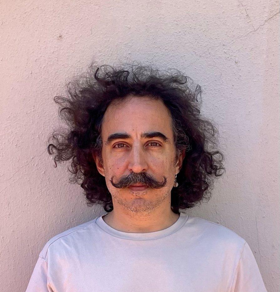 Retrato de Pablo Katchadjian, escritor argentino, frente a pared clara con cabello rizado y bigote