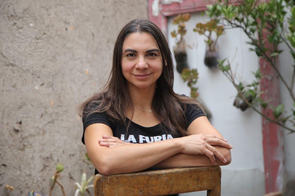 Mujer joven con cabello oscuro, brazos cruzados, sonriendo hacia la cámara frente a pared de cemento con plantas