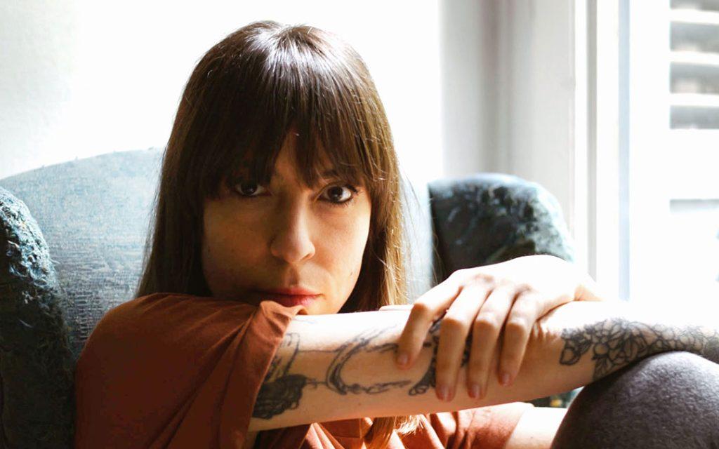 Mujer con cabello negro y flequillo, apoyada en su brazo junto a una ventana con vista urbana