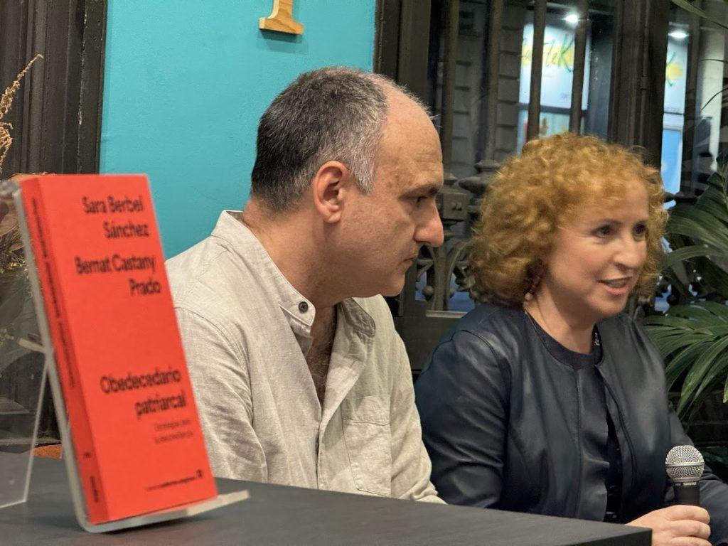 Dos personas en una mesa de panel: hombre de mediana edad con camisa beige y mujer rubia con blazer negro, frente