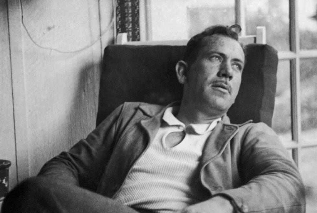 John Steinbeck sentado en una silla, mirando hacia arriba pensativo, en blanco y negro