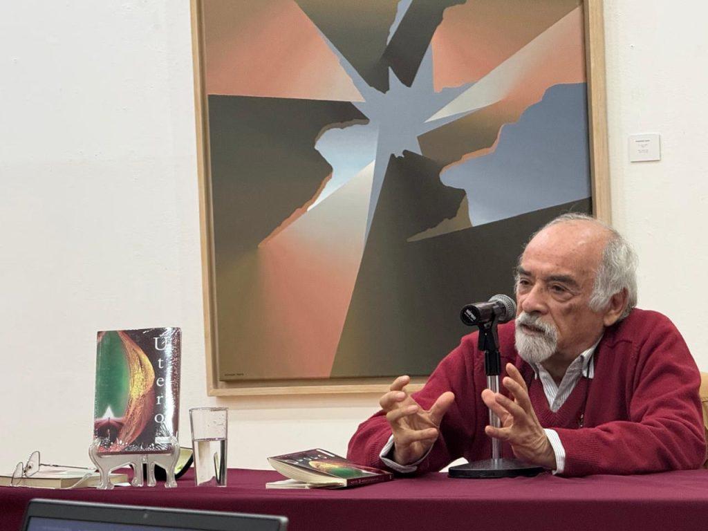 Juan Mihovilovich durante una conferencia, gesticulando mientras habla ante micrófono, con cuadro abstracto al fondo