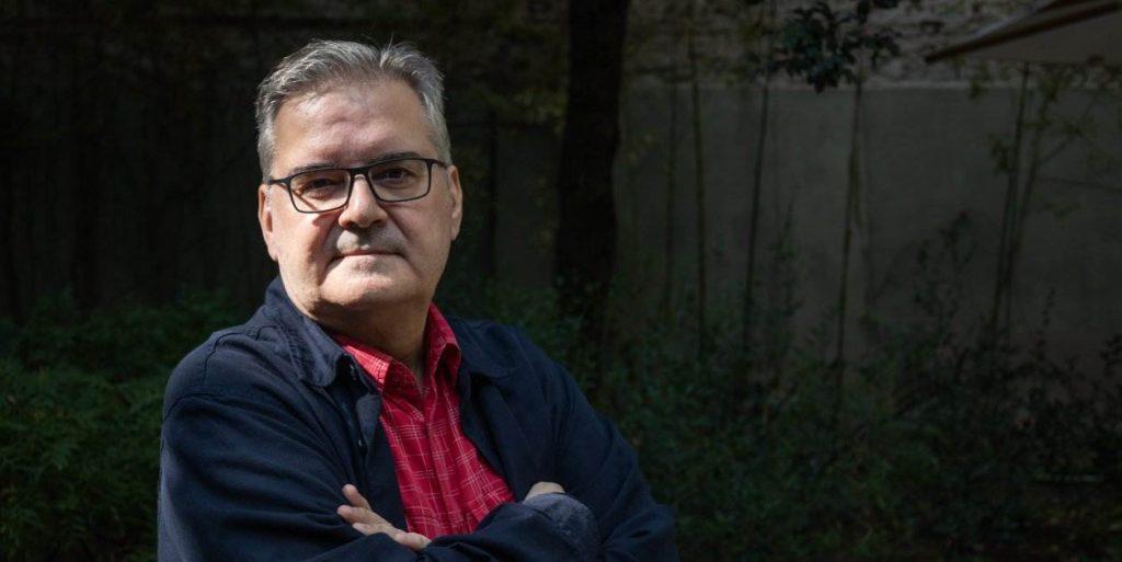 Sergi Pàmies, escritor, retratado con gafas y suéter azul marino sobre fondo de vegetación