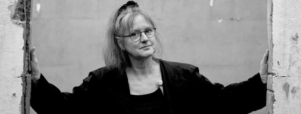 Solvej Balle con gafas, cabello largo y chaqueta negra, posando con brazos extendidos en fotografía en blanco y negro