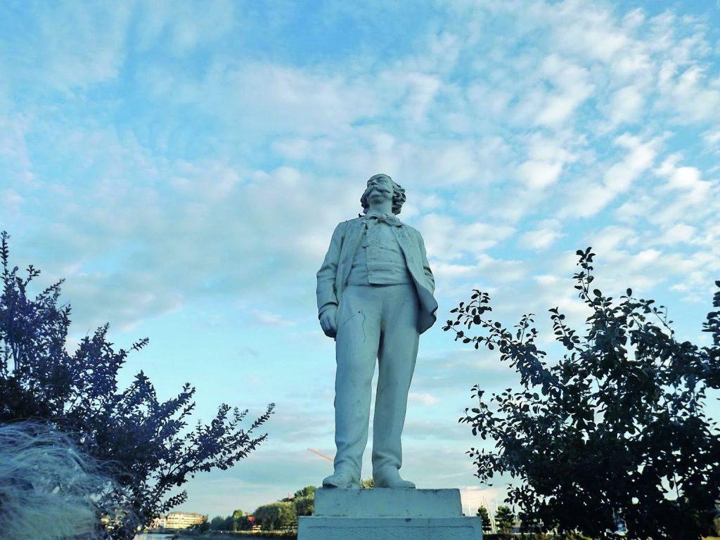 Estatua de Gustave Flaubert de pie sobre pedestal, rodeada de vegetación, contra cielo azul con nubes