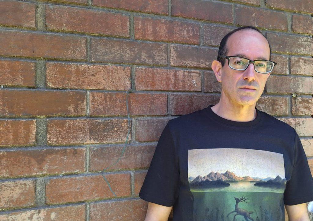 Nicolás Poblete Pardo, escritor, posa frente a pared de ladrillo rojo vistiendo camiseta negra con estampado de paisaje