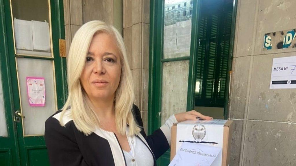 Mujer de cabello rubio platinado, blusa blanca y chaqueta negra, frente a puerta verde en interior de edificio histórico