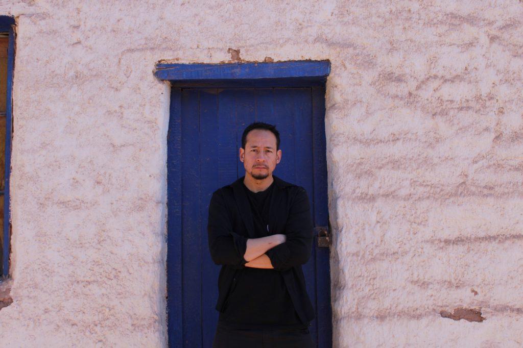 Hombre de camiseta negra posando frente a puerta azul en pared rosada, brazos cruzados