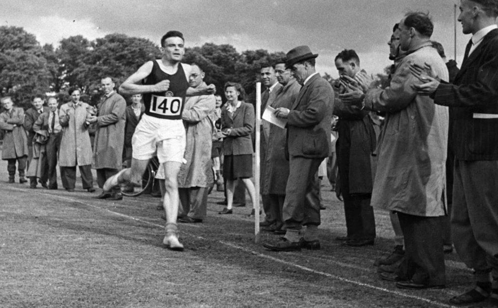 Alan Turing corre en una competencia de atletismo rodeado de espectadores en un evento deportivo de mediados del siglo