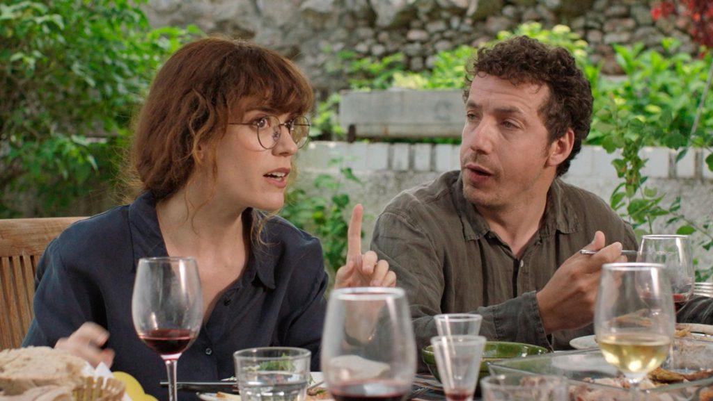 Dos personajes conversan en una mesa durante una cena en ambiente exterior con vegetación de fondo