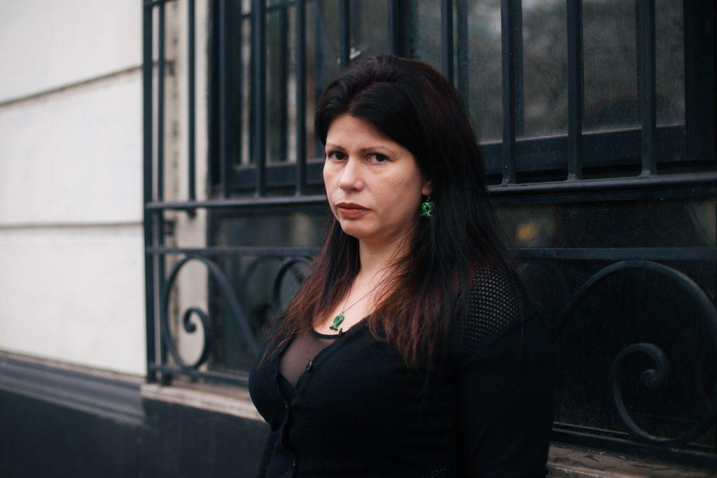 Mujer de cabello oscuro largo con blazer negro, posada frente a puerta oscura en ambiente urbano