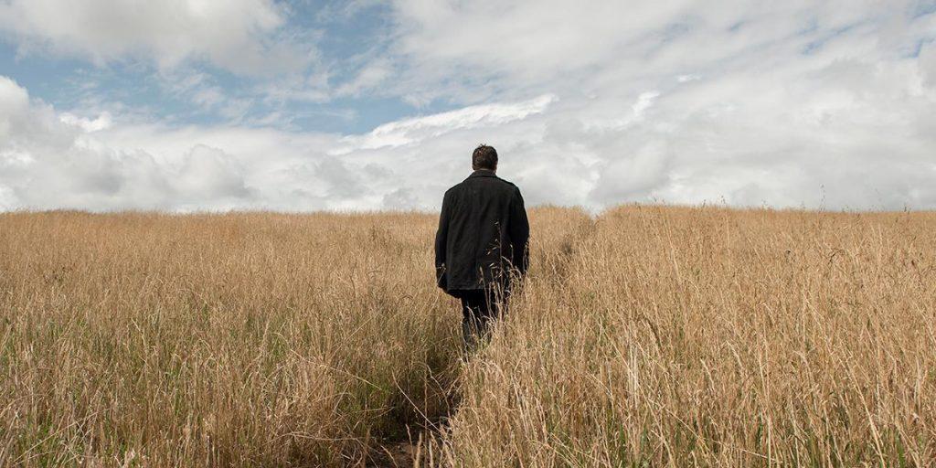 Hombre de espaldas caminando solo por un vasto campo de trigo seco bajo cielo nublado