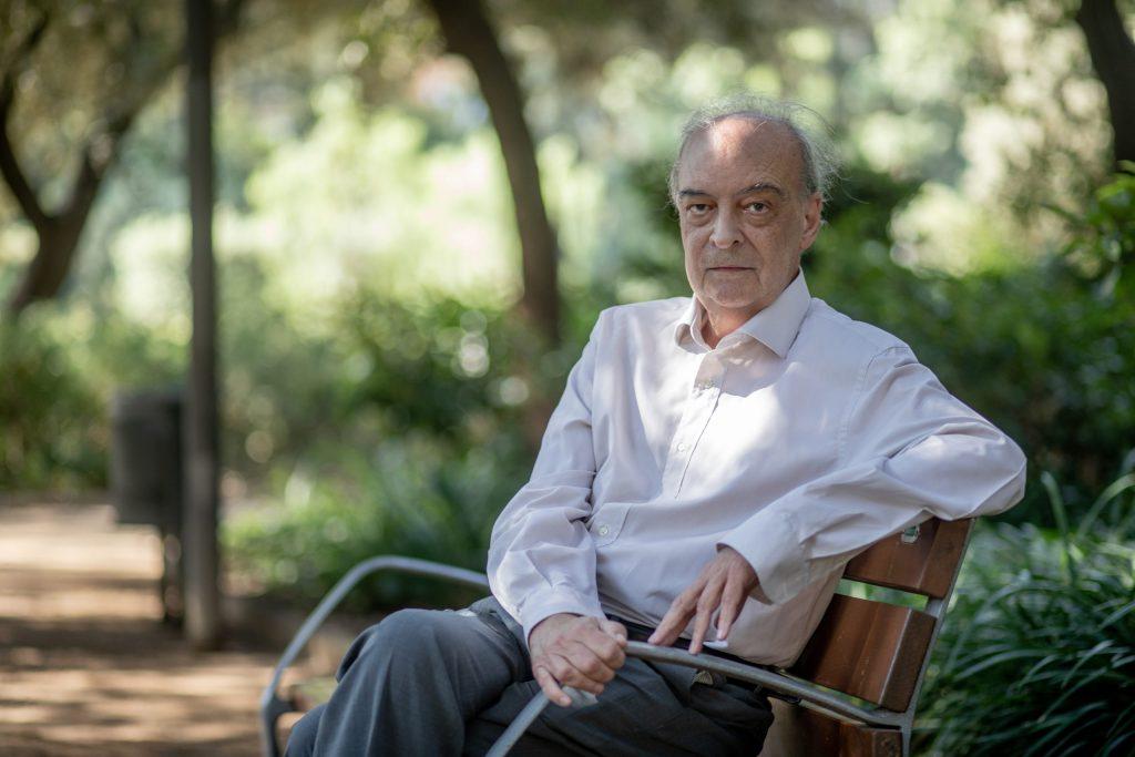 Enrique Vila-Matas sentado en un banco de parque sosteniendo un libro, con árboles de fondo