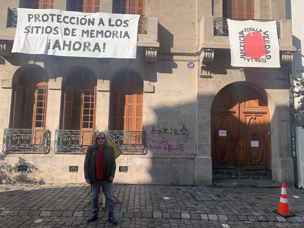 Fachada de edificio histórico con persona frente a puerta de madera, cartel de protección a sitios de memoria y símbolo