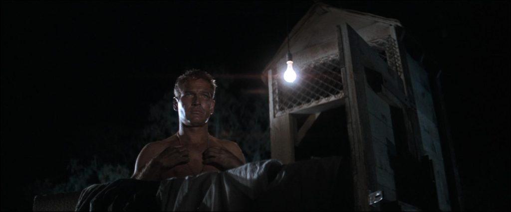 Hombre en traje sosteniendo una puerta de metal iluminada en la oscuridad, escena de tensión dramática