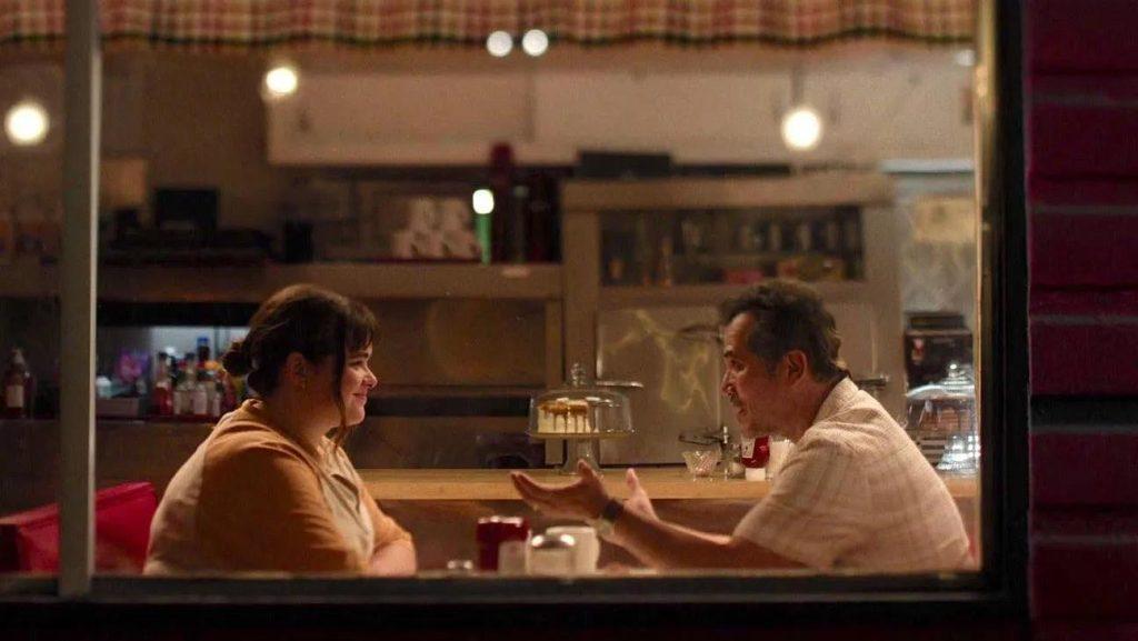 Dos hombres en una cafetería por la noche, sentados frente a frente en una mesa, conversando intensamente mientras