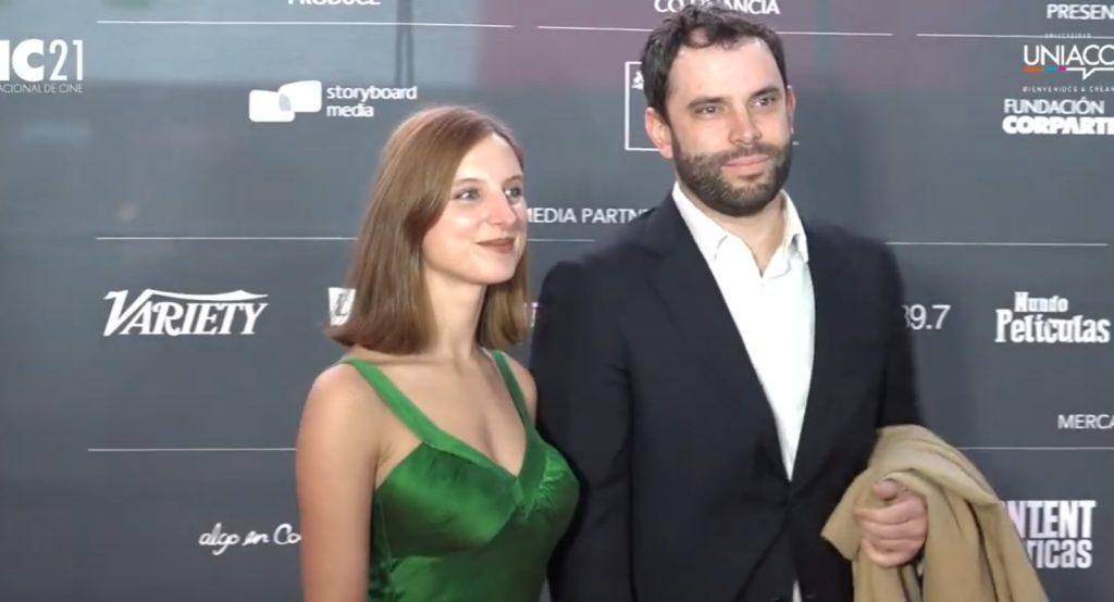 Pareja en alfombra roja de evento cultural: mujer con vestido verde esmeralda y hombre de traje negro posando juntos