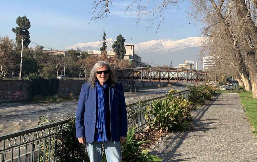 Mujer con abrigo azul marino y bufanda frente a puente y iglesia con montañas nevadas al fondo en ciudad andina