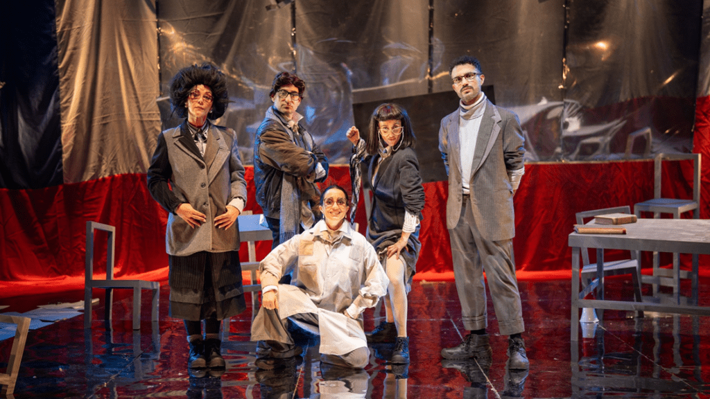 Escena teatral con cinco actores en un escenario rojo; uno sentado al frente en camisa blanca, otros de pie al fondo