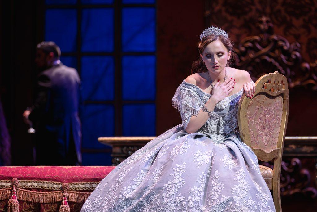 Mujer en vestido blanco elegante sentada en sillón dorado durante escena teatral de La traviata, con actor de fondo