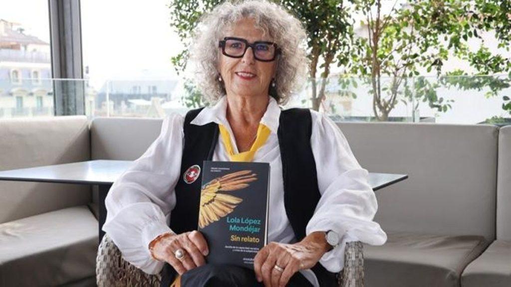 Mujer mayor con cabello blanco y gafas sostiene libro de tapa amarilla, sentada en sofá gris junto a ventana