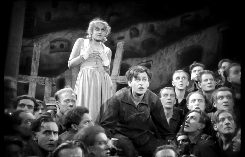 Escena de cine mudo en blanco y negro: mujer en vestido claro de pie entre multitud de personas, ambiente urbano