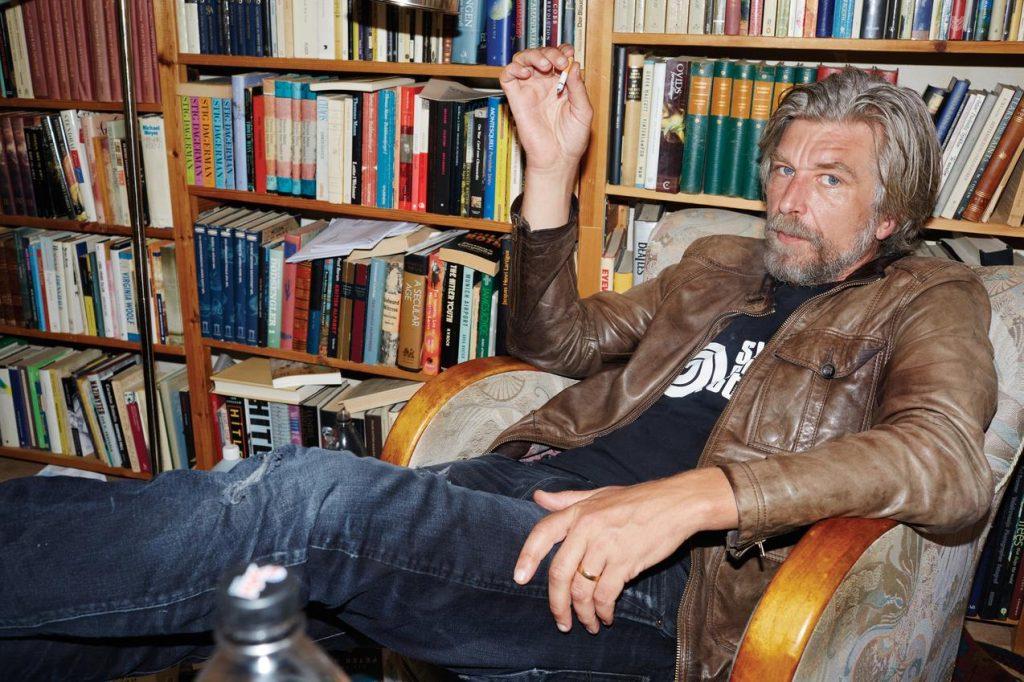 Hombre con barba recostado en sillón frente a estantería llena de libros, mirando hacia un lado