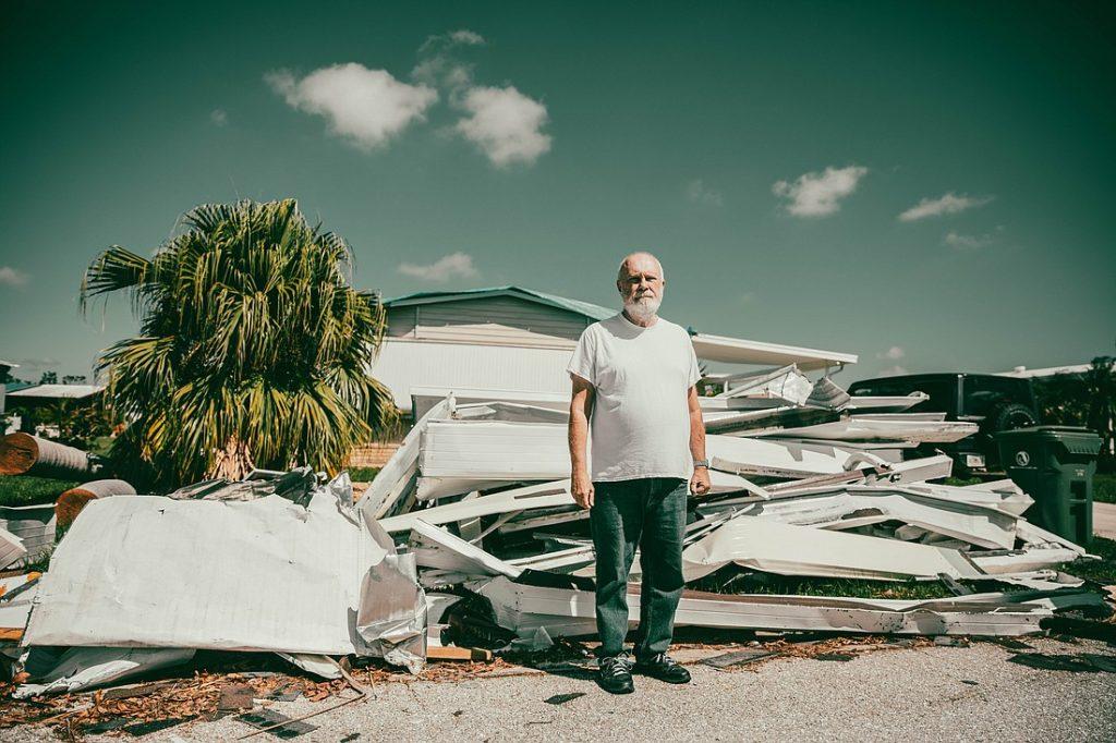 Hombre de cabello blanco y gafas de sol de pie entre escombros y materiales de desecho en un terreno abandonado