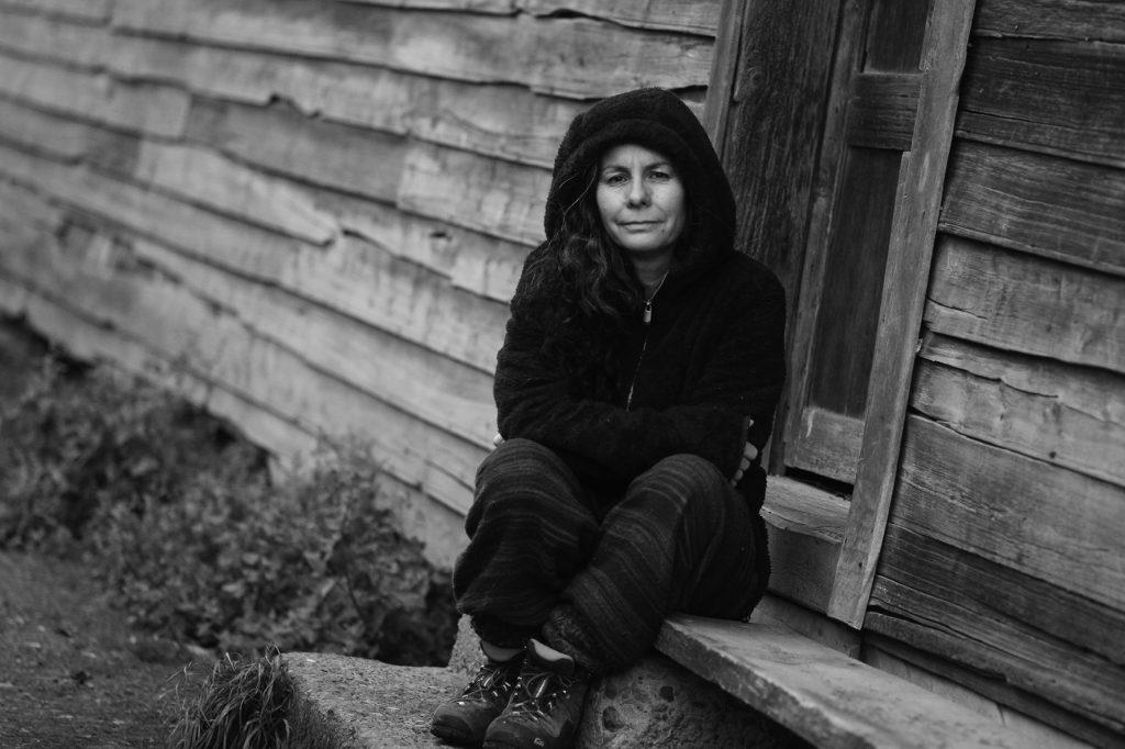 Poeta Verónica Quense sentada contra pared de madera, con capucha negra, en fotografía blanco y negro