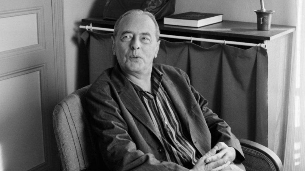 Witold Gombrowicz, escritor polaco, sentado en una silla frente a una estantería en blanco y negro
