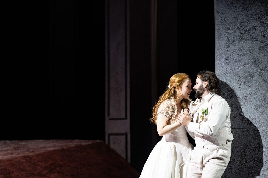 Escena teatral: mujer de cabello rubio en vestido blanco abraza a hombre de cabello oscuro en traje claro bajo luz