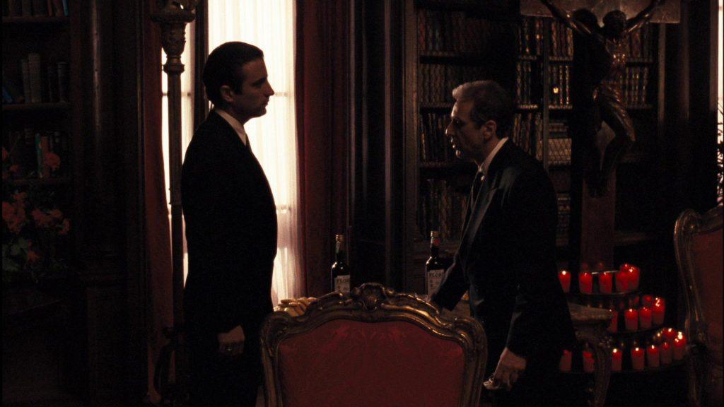 Dos hombres en traje conversan en interior oscuro con velas encendidas de fondo, escena de drama político y criminal