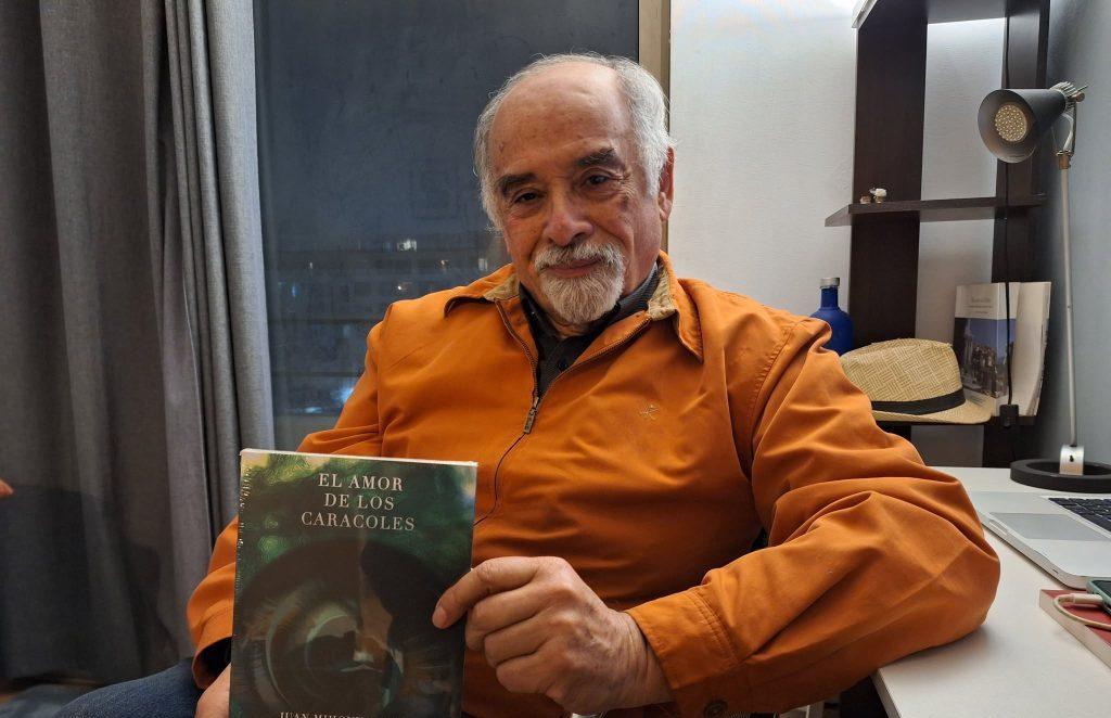 Hombre mayor con cabello blanco vistiendo chaqueta naranja sostiene un libro de portada oscura en interior