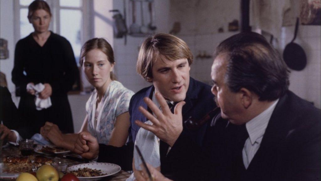 Cuatro personas en una cocina durante una conversación tensa; hombre de traje oscuro gesticula mientras habla con otro