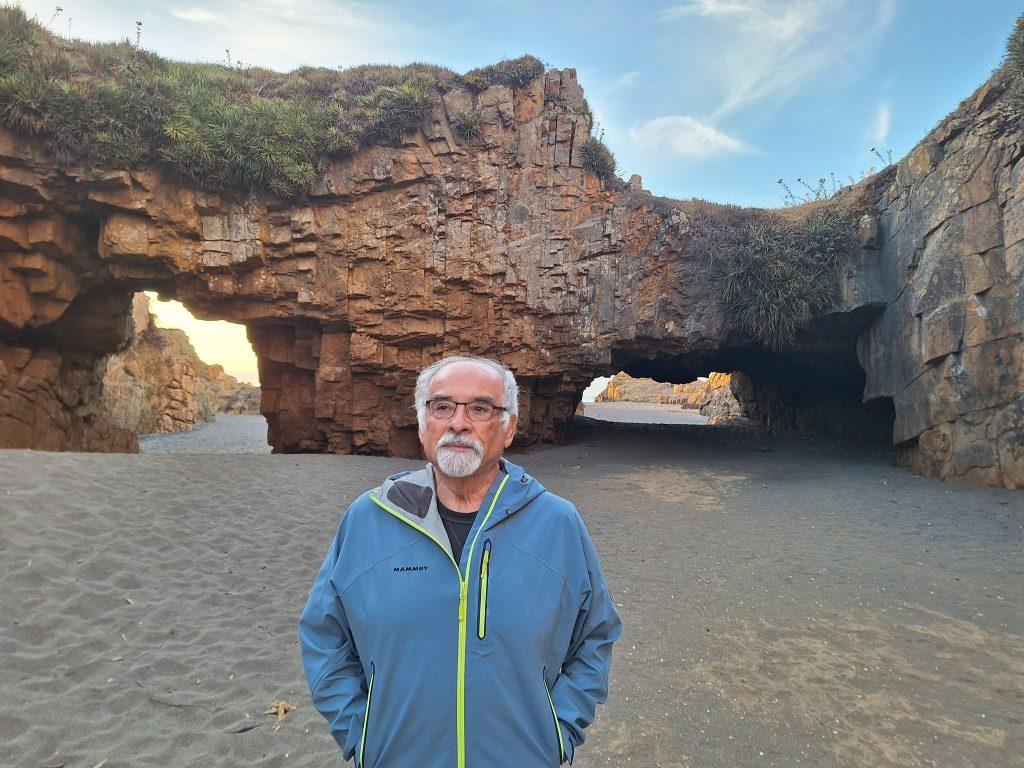 Hombre con gafas y chaqueta azul ante formaciones rocosas con arcos naturales en playa