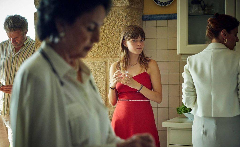 Escena de interior en casa antigua. Mujer en vestido rojo de pie en primer plano, hombres en ropa clara conversando